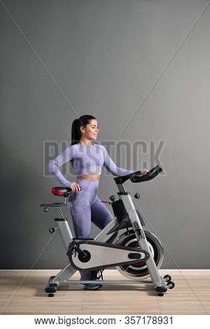 A Beautiful Athletic Young Brunette Woman In Sportswear Trains On A Sycle In The Gym Against The Gra