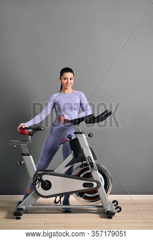 A Beautiful Athletic Young Brunette Woman In Sportswear Trains On A Sycle In The Gym Against The Gra
