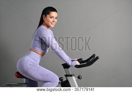 A Beautiful Athletic Young Brunette Woman In Sportswear Trains On A Sycle In The Gym Against The Gra