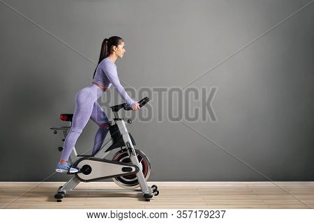 A Beautiful Athletic Young Brunette Woman In Sportswear Trains On A Sycle In The Gym Against The Gra