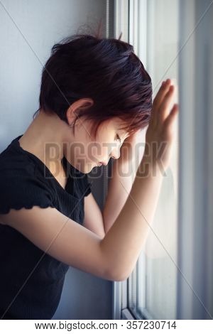 Vertical Picture Of Sad Little Girl Looking Out Window Touching Glass By Hand. Child During Quaranti