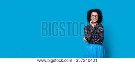 Cheerful Caucasian Lady With Curly Hair Smiling While Touching Her Chin On A Blue Background With Fr