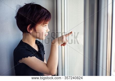 Sad Little Girl Looking Out Window Touching Glass By Hand, Pointing Finger. Child During Quarantine