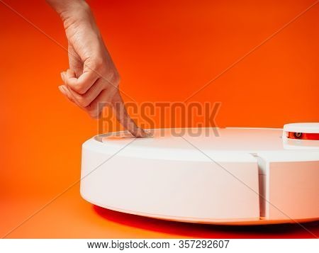 Girl Presses A Hand The Power Button On A Robot Vacuum Cleaner On An Orange Background.