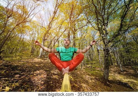 An Adult Bearded Barefoot Man Sits And Meditates On A Slackline In The Autumn Forest. Yoga And Balan