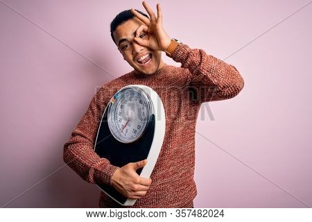 Young brazilian man doing diet to lose weigth holding scale over isolated pink background with happy face smiling doing ok sign with hand on eye looking through fingers