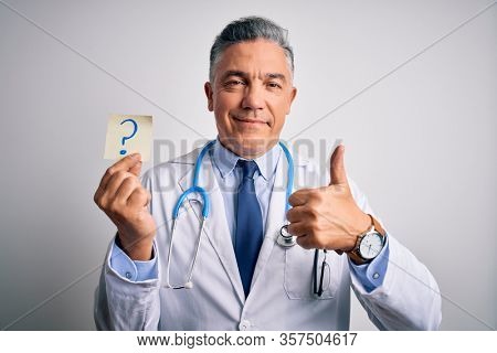 Middle age handsome grey-haired doctor man holding paper with question mark happy with big smile doing ok sign, thumb up with fingers, excellent sign