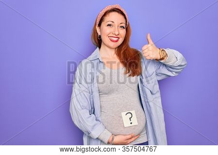 Young beautiful redhead pregnant woman expecting baby with question mark on stomach happy with big smile doing ok sign, thumb up with fingers, excellent sign