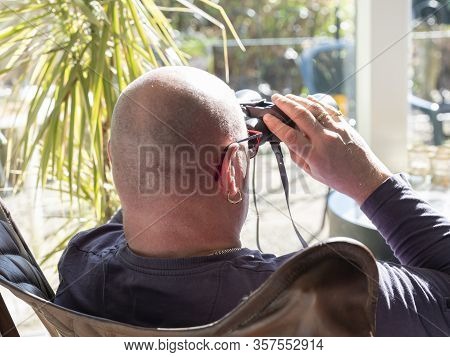 Elderly Man Sittiing, Looking Through A Glass Door With Binoculars As He Watches Something In The Di