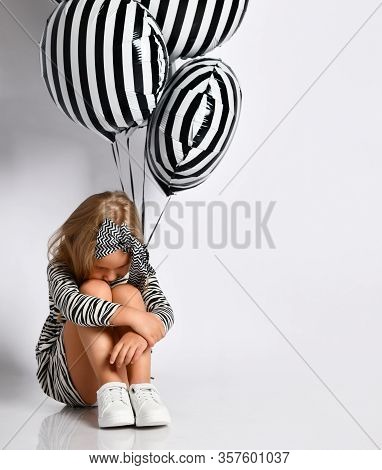 Child In Dress, Headband And Sneakers. She Put Her Arms Around Legs And Looking Sad, Holding Striped
