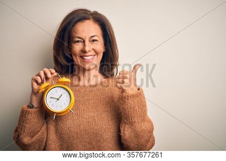 Middle age brunette woman holding clasic alarm clock over isolated background Pointing to the back behind with hand and thumbs up, smiling confident