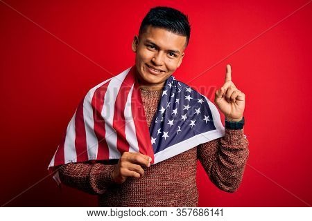 Young handsome latin patriotic man wearing united states of america falg surprised with an idea or question pointing finger with happy face, number one