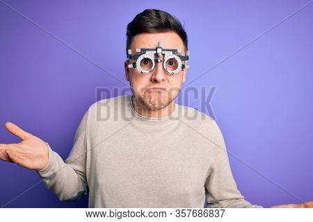 Young handsome caucasian man wearing optometrical glasses over purple background clueless and confused expression with arms and hands raised. Doubt concept.