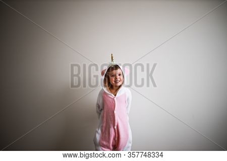 Little Todder Girl Standing Next To A White Wall Wearing Unicorn Kigurumi And Looking At Camera