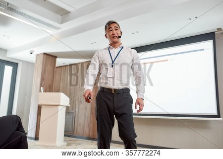 Asian Male Speaker In Suit Giving A Talk At Corporate Business Meeting, Forum. Chart On The Screen I
