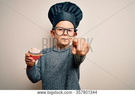 Young little caucasian cook kid wearing chef uniform and hat cooking cupcake pointing with finger to the camera and to you, hand sign, positive and confident gesture from the front