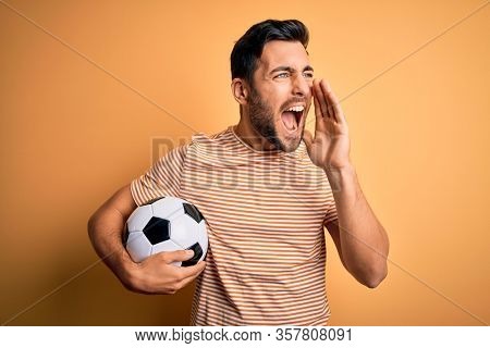 Handsome player man with beard playing soccer holding footballl ball over yellow background shouting and screaming loud to side with hand on mouth. Communication concept.