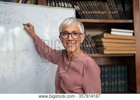 Portrait of happy mature professor teaching mathematics to students in a library. Senior smiling woman solving math problem while writing on white board. Portrait of tutor looking at camera.