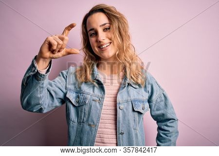 Young beautiful blonde woman wearing casual denim jacket standing over pink background smiling and confident gesturing with hand doing small size sign with fingers looking and the camera. Measure