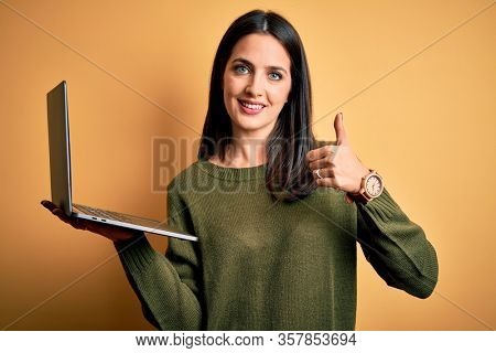 Young brunette woman with blue eyes working using computer laptop over yellow background doing happy thumbs up gesture with hand. Approving expression looking at the camera showing success.