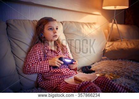 Happy Little Girl In Pajams Sitting On Sofa, Eating Pizza And Playing Video Games With Joystick At H