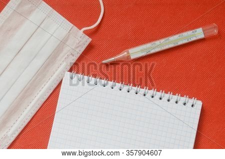Top View, Medical Mask, Termometer And Emty Notepad On Red Background.
