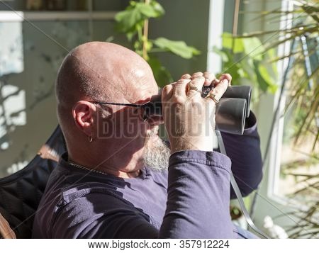Elderly Man Sittiing, Looking Through A Glass Door With Binoculars As He Watches Something In The Di