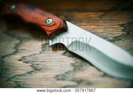 A Sharp Steel Hunting Knife With A Wooden Handle Lies On A Wooden Table, Illuminated By The Light.