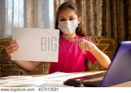 Young Schoolgirl In Medical Mask Pointing To Blank Cheet Of Paper For Inscription. Concept Of Distan