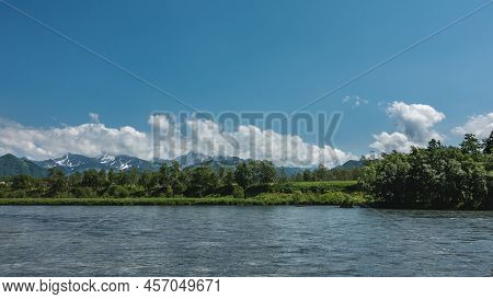 Lush Green Vegetation Grows On The Bank Of A Calm Blue River - Grass, Trees. A Mountain Range Agains