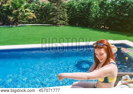 Young Girl Sunbathing By The Pool, Sitting On An Amaca Applying Sun Cream On Her Arms. Woman Enjoyin