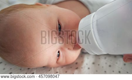 Newborn Baby Girl With Half Closed Eyes Drinks Water From Plastic Bottle Lying On Bed. Mother Feeds 