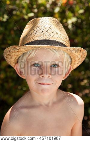 Portrait Of The Blode Hair Boy Smiling With Blue Eyes And Straw Hat With Blurred Summer Greenery Bac
