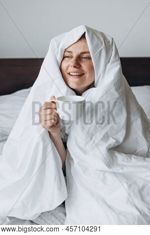 Young Happy Redhead Woman With Cup Of Coffee Sitting In Bed Under Blanket At Home Bedroom. Comfort O