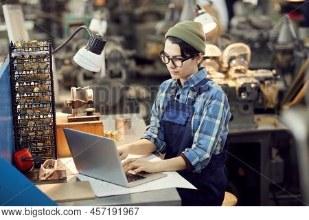 Concentrated Hipster Girl In Beanie Hat Sitting At Table In Watch Factory Shop And Using Laptop Whil