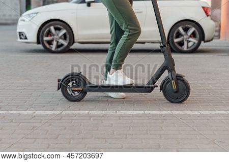 Young Woman In White Sneakers Is Standing On Electric Scooter In Front Of The White Car