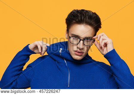 An Attractive Young Man Stands On An Orange Background In A Blue Zip-up Jacket And Black Eyeglasses 