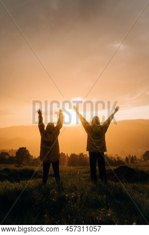 Two Hikers In Raincoats Stand In The Mountains At Sunset In The Rain With Their Hands Up And Look At