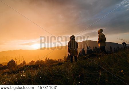 Two Hikers Stand In The Rain On A Meadow In The Mountains And Watch The Incredible Orange Sunset. Tw