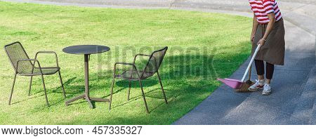 Female Worker Sweeping Yard With Broom Tool And Dustpan In Ornamental Backyard Garden. Woman Wear Ap