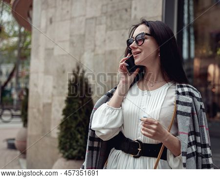 Attractive Cheerful Young Woman In Sunglasses Speaking On Cellphone In Town Street And Smiling.