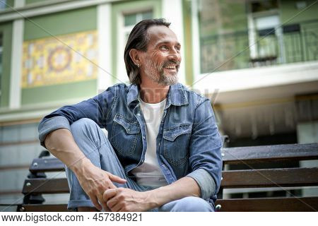 Handsome Happy Smiling Middle Aged Man With Grey Beard Sits On The Bench With Hands Leaned On Lifted