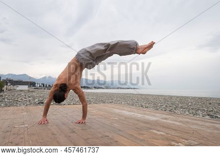 Shirtless Caucasian Man Doing Backflip On Pebble Beach.