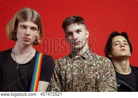 Three Young Handsome Men Of Different Nationalities Stand On Vibrant Red Background. Gay Relationshi