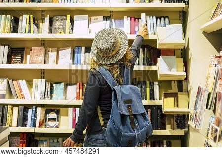 Back View Of Woman With Hat And Backpack Choosing A Book To Buy Inside A Library Or Newspaper Store.