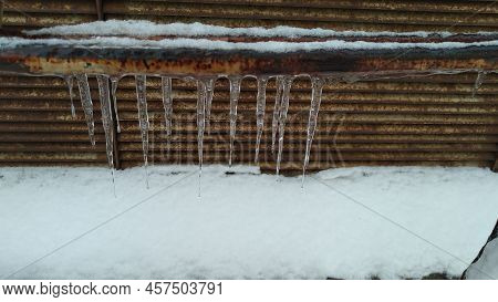 Several Ice Icicles Hang From An Iron Pipe. A Rusty Iron Pipe Covered With Seng, With Icicles Hangin