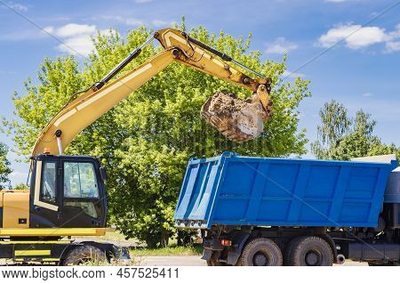 An Excavator Against A Blue Sky Loads Excavated Soil Into A Dump Truck. Loading Earth Into A Car Usi