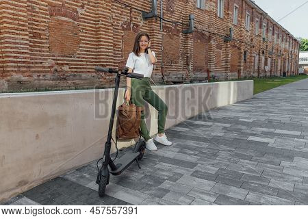 Woman In White Sneakers And Leather Backpack Standing Near Electric Scooter