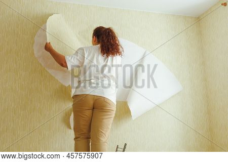 Back View Of Buxom Woman Wearing White T-shirt, Beige Trousers, Standing On Chair, Tearing Off Old W