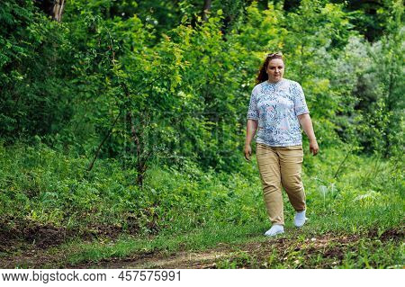 Side View Of Young Plump Buxom Woman With Long Curly Red Hair Walking On Path In Park Forest Among G